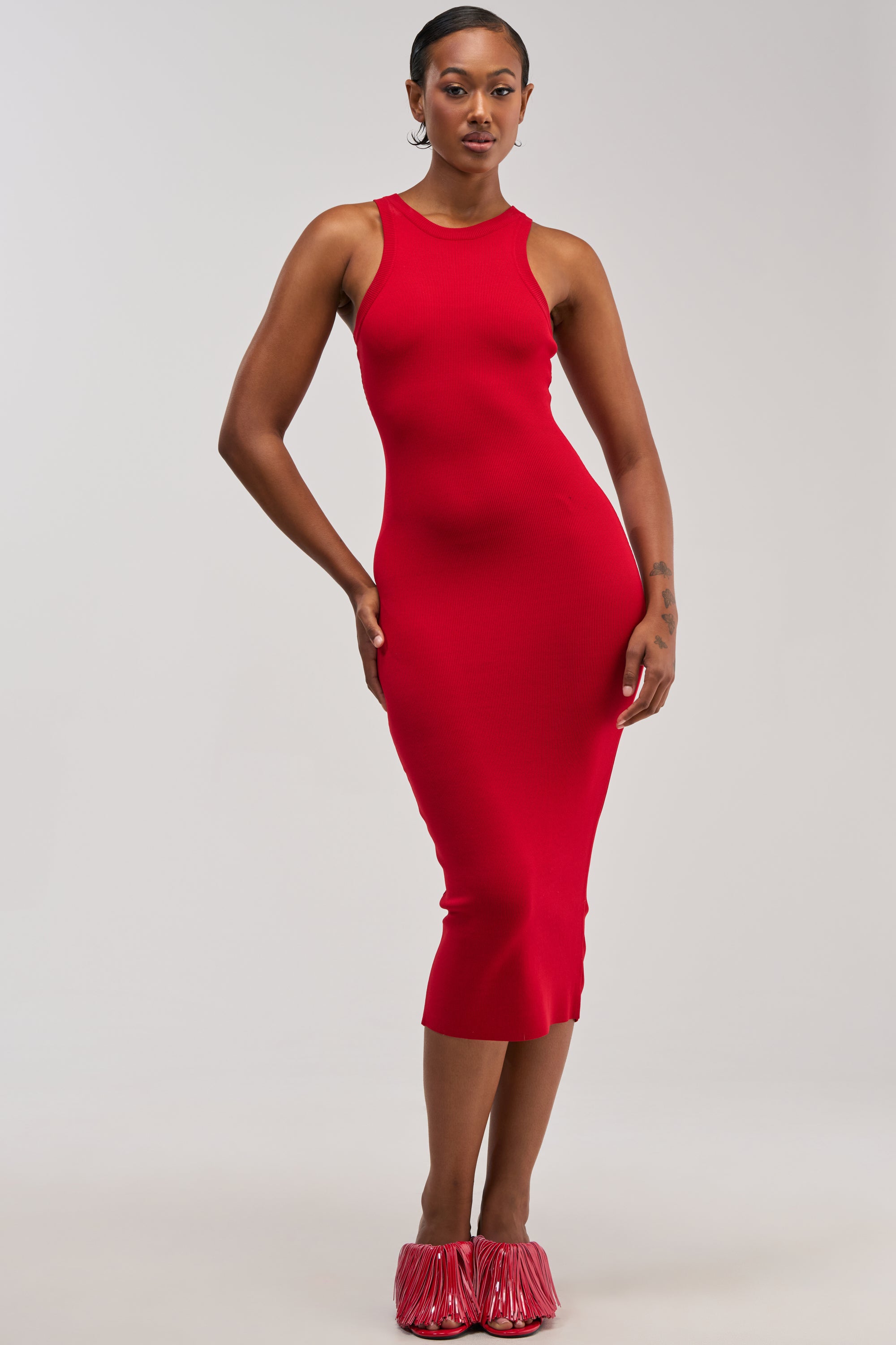 A person stands confidently wearing the CUTIE PIE KNIT MIDI DRESS IN RED, a fitted, sleeveless knit tank dress styled for summer, paired with red fringe-detailed shoes against a minimal light gray background.
