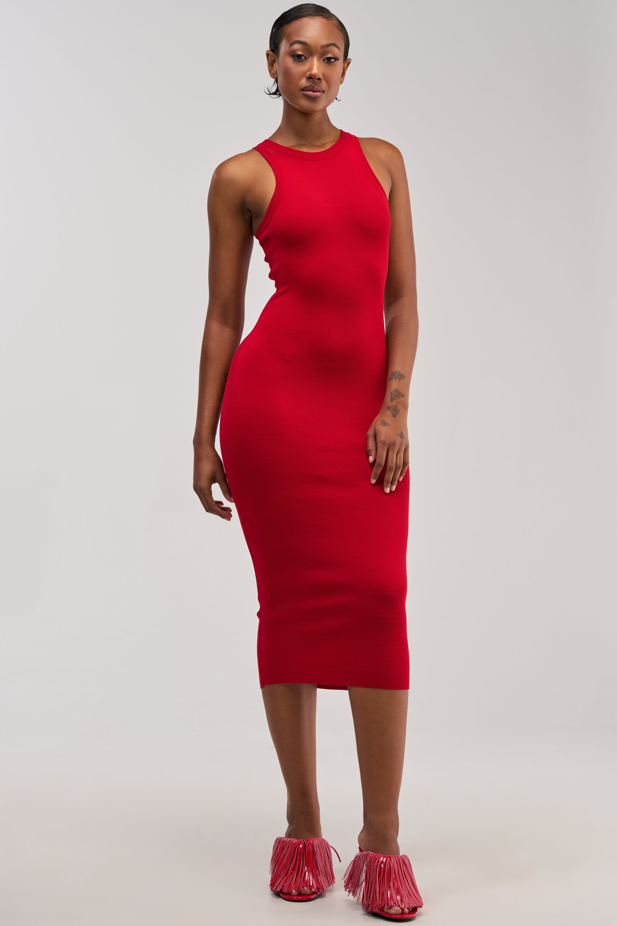 A woman with short hair stands against a plain background, wearing the CUTIE PIE KNIT MIDI DRESS IN RED. The sleeveless, form-fitting knit dress is styled for an effortless summer look, paired with red fringe slip-on shoes as she poses with one arm relaxed by her side.