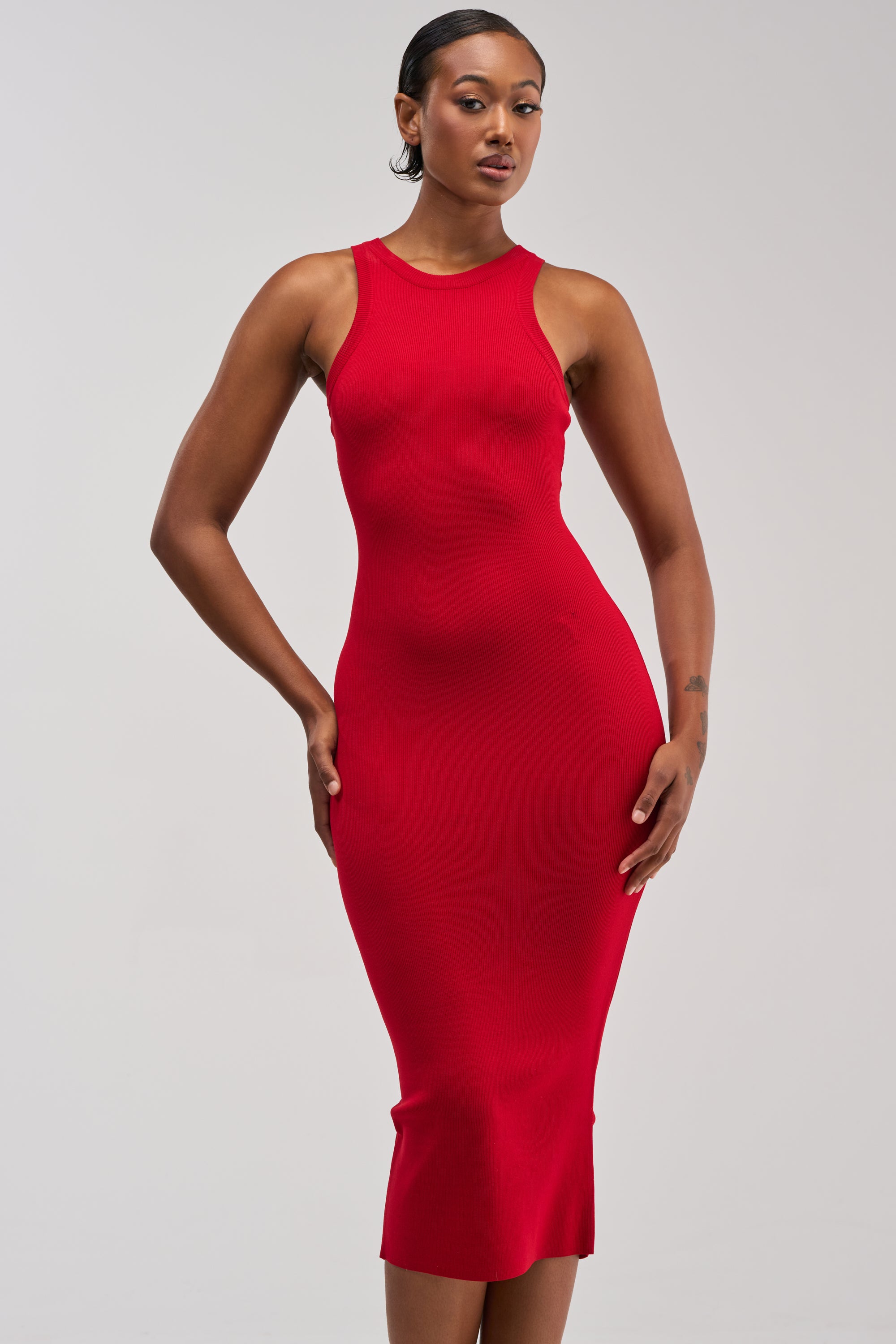 A woman models the CUTIE PIE KNIT MIDI DRESS IN RED—a sleeveless, fitted knit tank dress—standing confidently against a plain light gray background with one hand on her hip, making it the perfect choice for effortless summer wear.