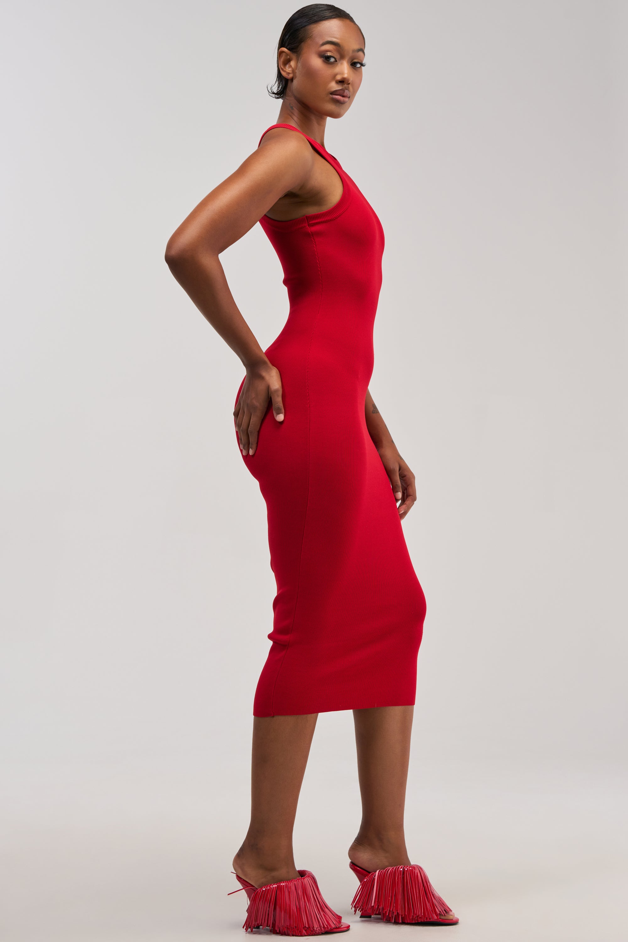 Wearing the CUTIE PIE KNIT MIDI DRESS IN RED, a woman stands sideways in red fringe heels, glancing toward the camera against a plain, light background—an effortlessly chic choice for summer wear.