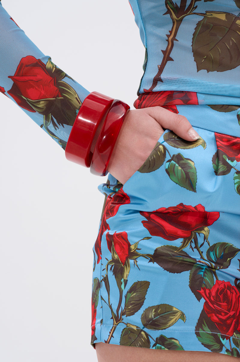 Model wearing the SUMMER TIME FINE BRACELET SET showcases chunky resin bangles on their wrist, paired with a blue outfit featuring a red rose pattern. One hand is in their pocket against a plain white background. 2