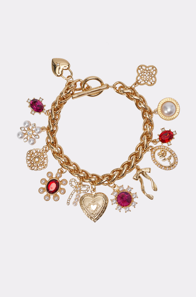 The CHARMED LIFE CHARM BRACELET features a chunky gold-tone chain adorned with dangling charms, including red gemstones, pearls, a flower, heart, shoe, and hand, displayed on a plain white background.