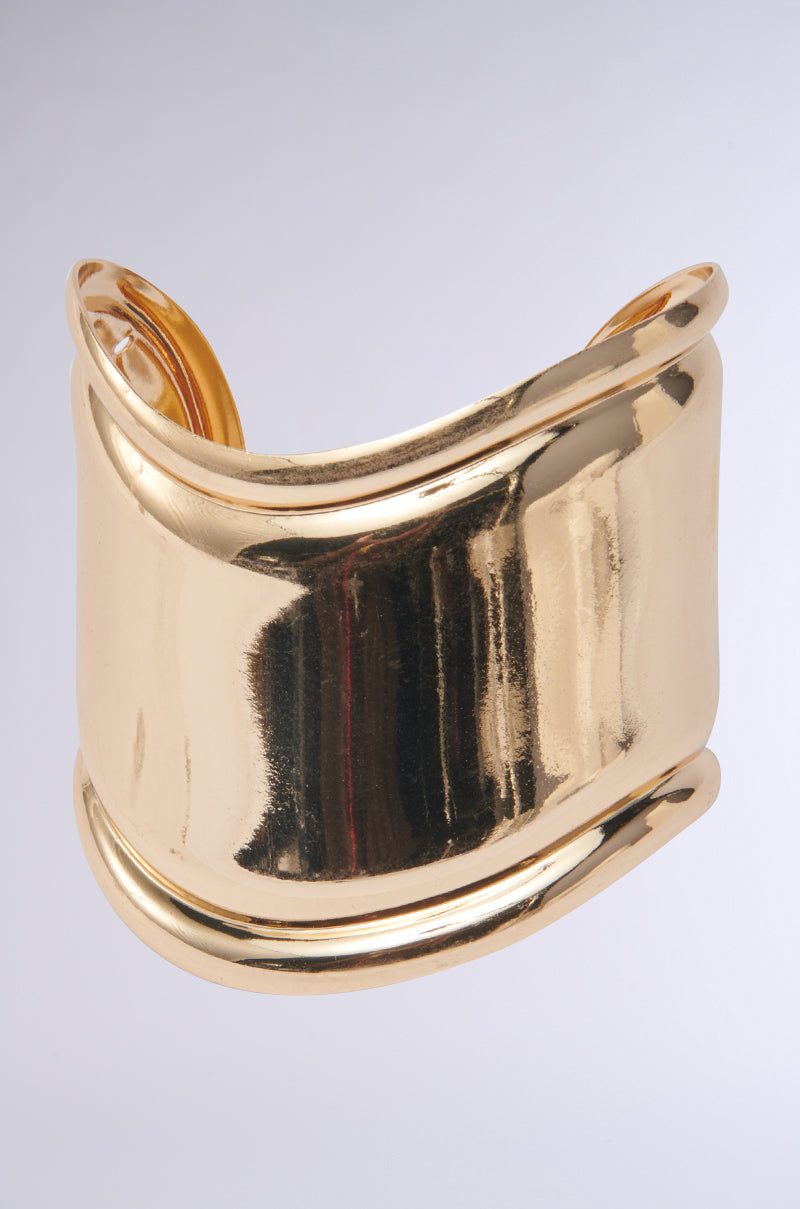 The IN THE MAGS BRACELET is a wide, shiny gold cuff with a smooth, reflective surface and gently curved edges, showcasing modern elegance against a plain light gray background.