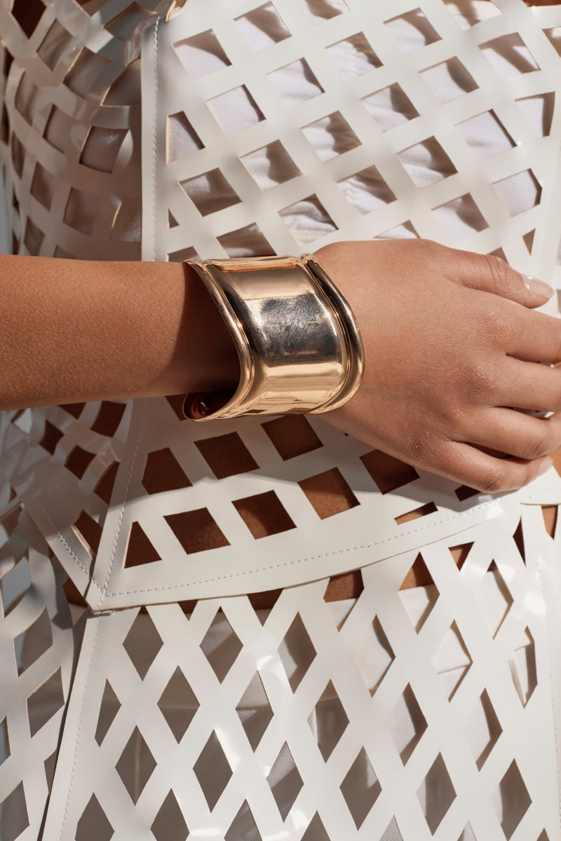 Wearing the IN THE MAGS BRACELET—a shiny gold sculptural cuff—this person radiates modern elegance, pairing it with a white geometric lattice-patterned outfit and striking a confident pose with hand on hip. 2