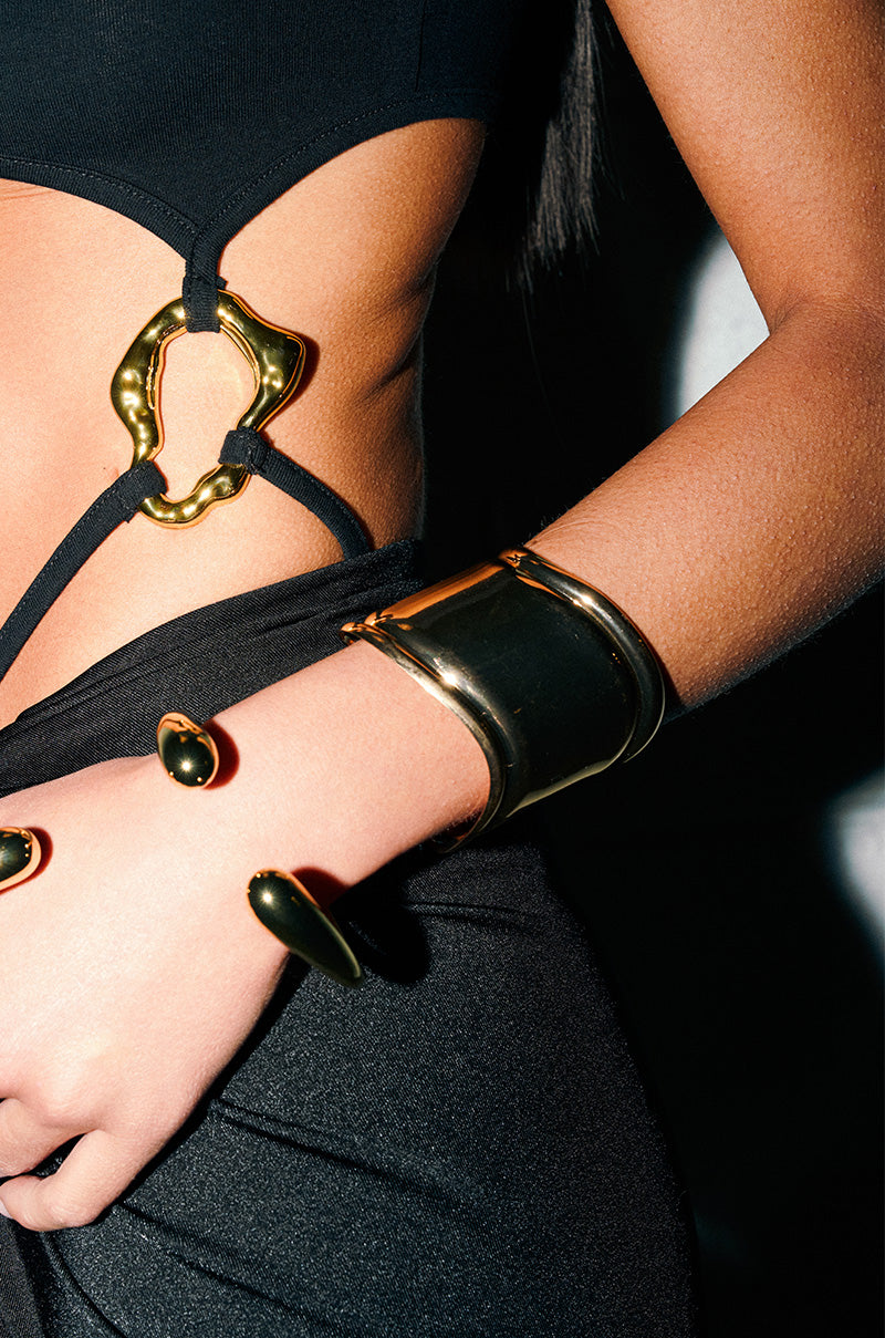 Against a neutral backdrop, a person in a modern black outfit showcases bold gold jewelry, including the sculptural IN THE MAGS BRACELET, plus a statement ring and a belt with striking gold hardware.