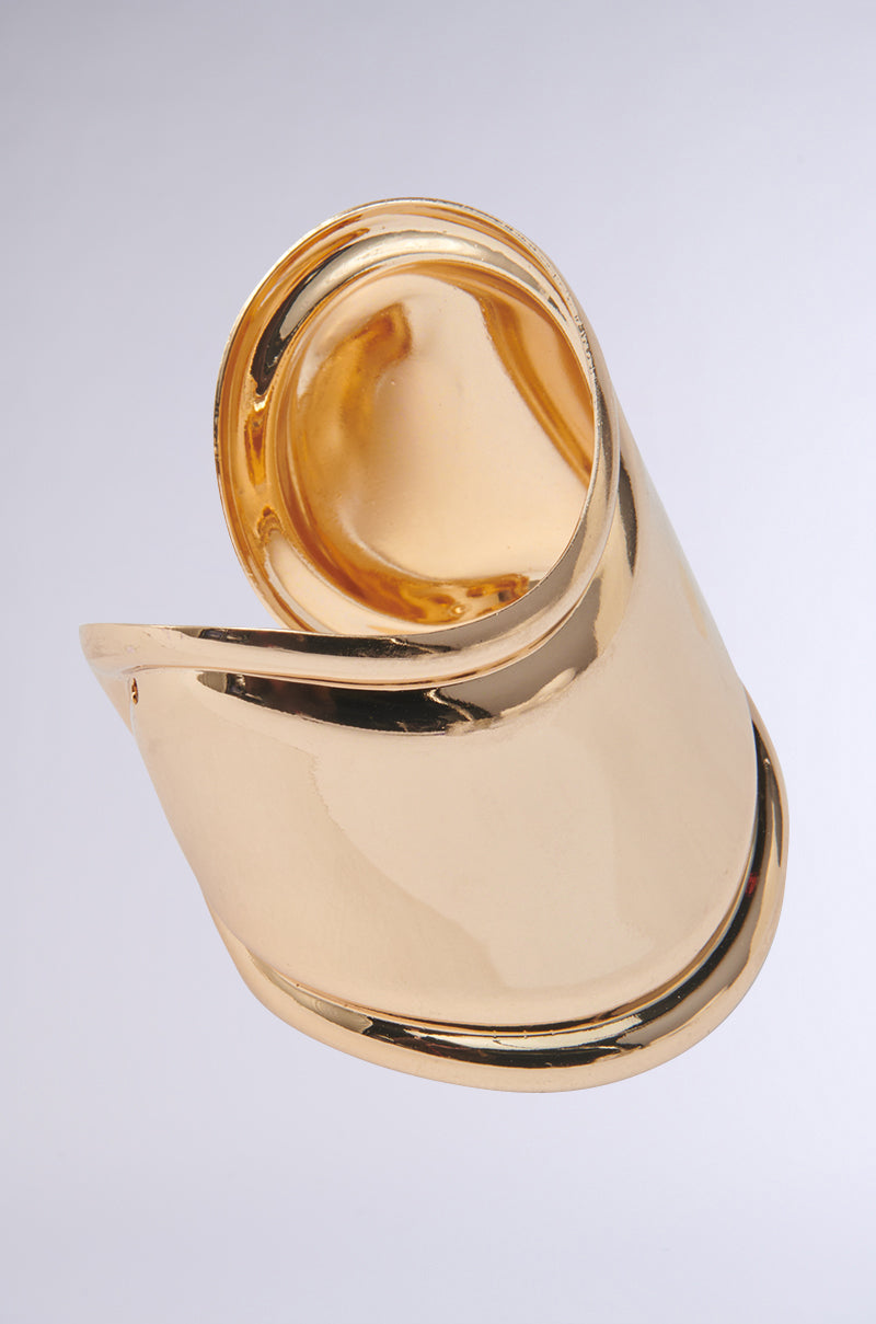 The IN THE MAGS BRACELET features a shiny gold, abstract sculptural design with smooth, curved surfaces and an open-ended style, showcasing modern elegance against a light gray background.