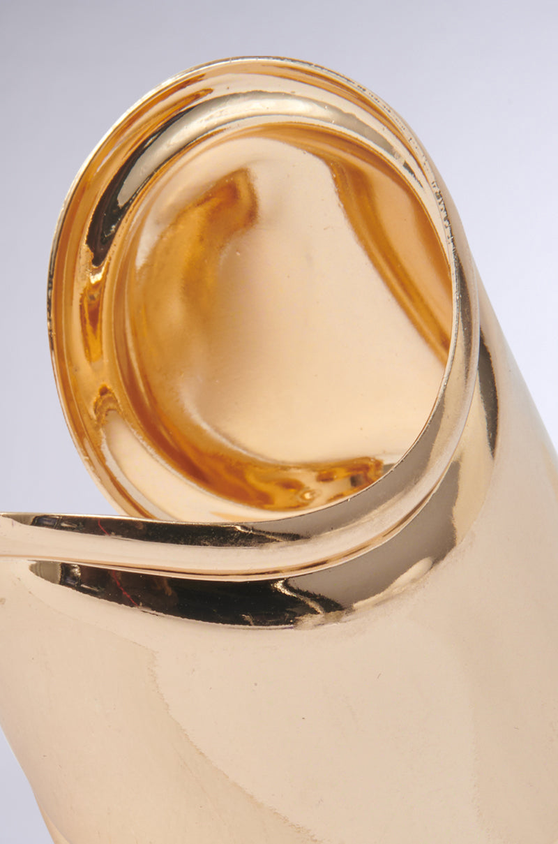 Close-up of the IN THE MAGS BRACELET—a shiny gold metallic piece with smooth, curved, rolled details that reflect light, showcasing a sculptural design and sleek modern elegance.