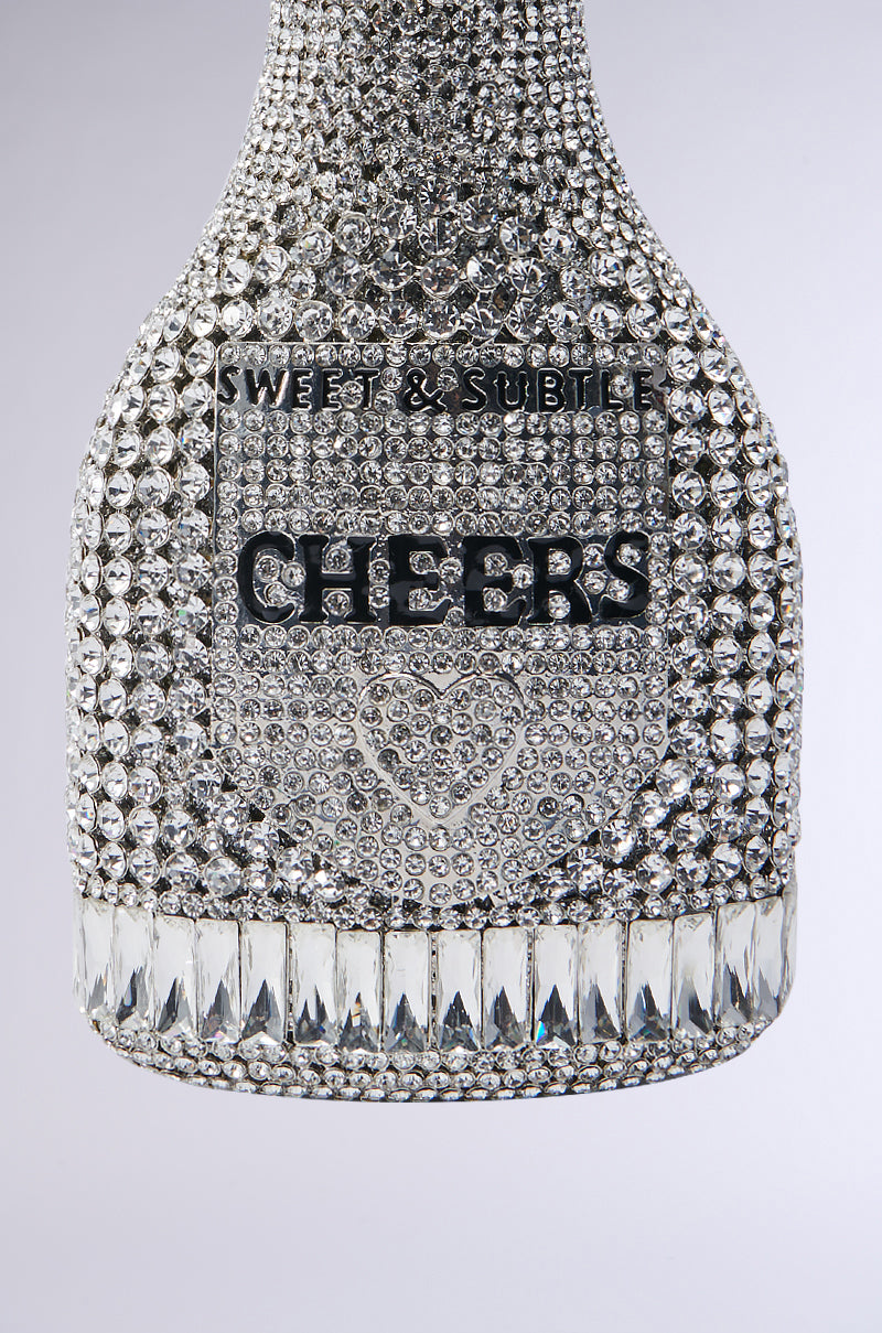 A close-up of the CHEERS BLING PURSE reveals its dazzling, rhinestone-covered surface featuring the words "Sweet & Subtle" and "Cheers." The base is adorned with larger, diamond-shaped silver rhinestones, complemented by a shimmering heart design underneath. Ideal for those who adore bling-inspired purse-style accessories. 3