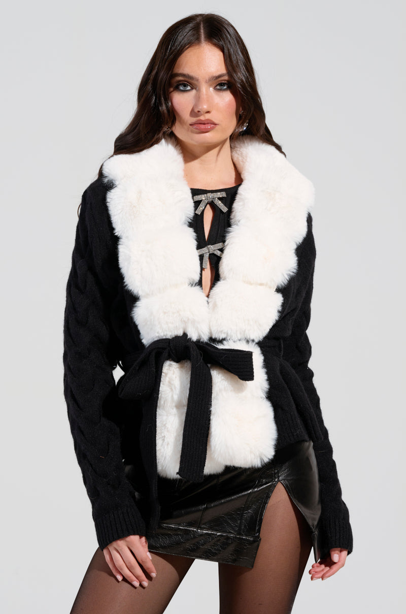 A woman with long dark hair is wearing the BABBS Knit Fur Trim Cardigan, characterized by its fur-trim collar and self-tie belt at the waist. She pairs it with a black leather skirt featuring a front slit and black tights, against a plain light gray background.