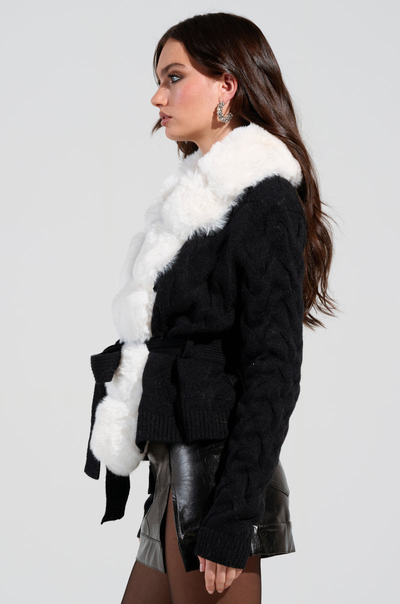 A woman with long brown hair is wearing the BABBS Knit Fur Trim Cardigan, featuring a white fur trim collar and a self-tie belt, along with a black leather skirt and matching tights, as she faces to the right against a light gray background.