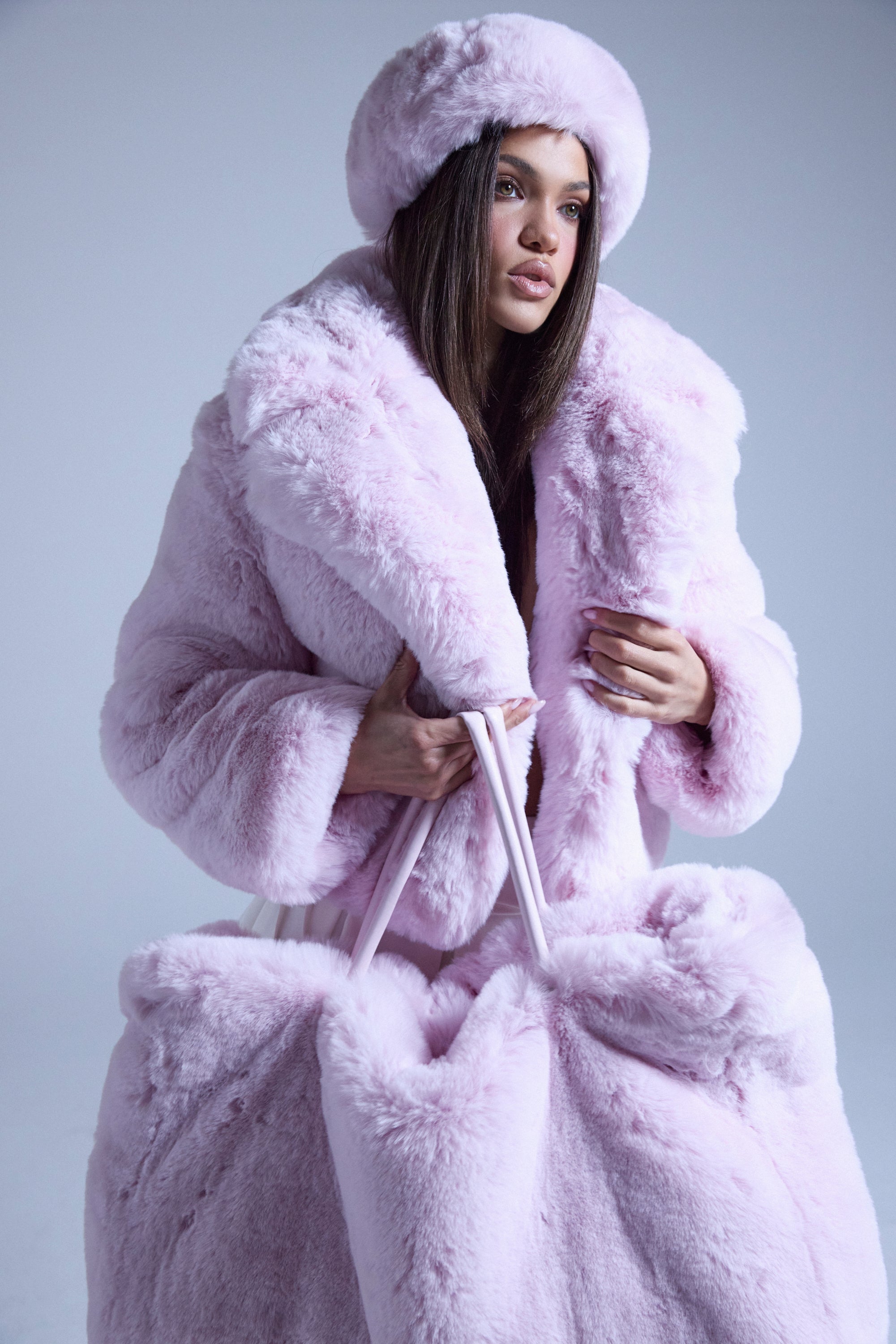 A woman wears the BBGIRL FAUX FUR COAT in fluffy light pink with an oversized collar and matching hat, holding a large fur bag. The plain background highlights the soft textures of her chic pink ensemble. 1