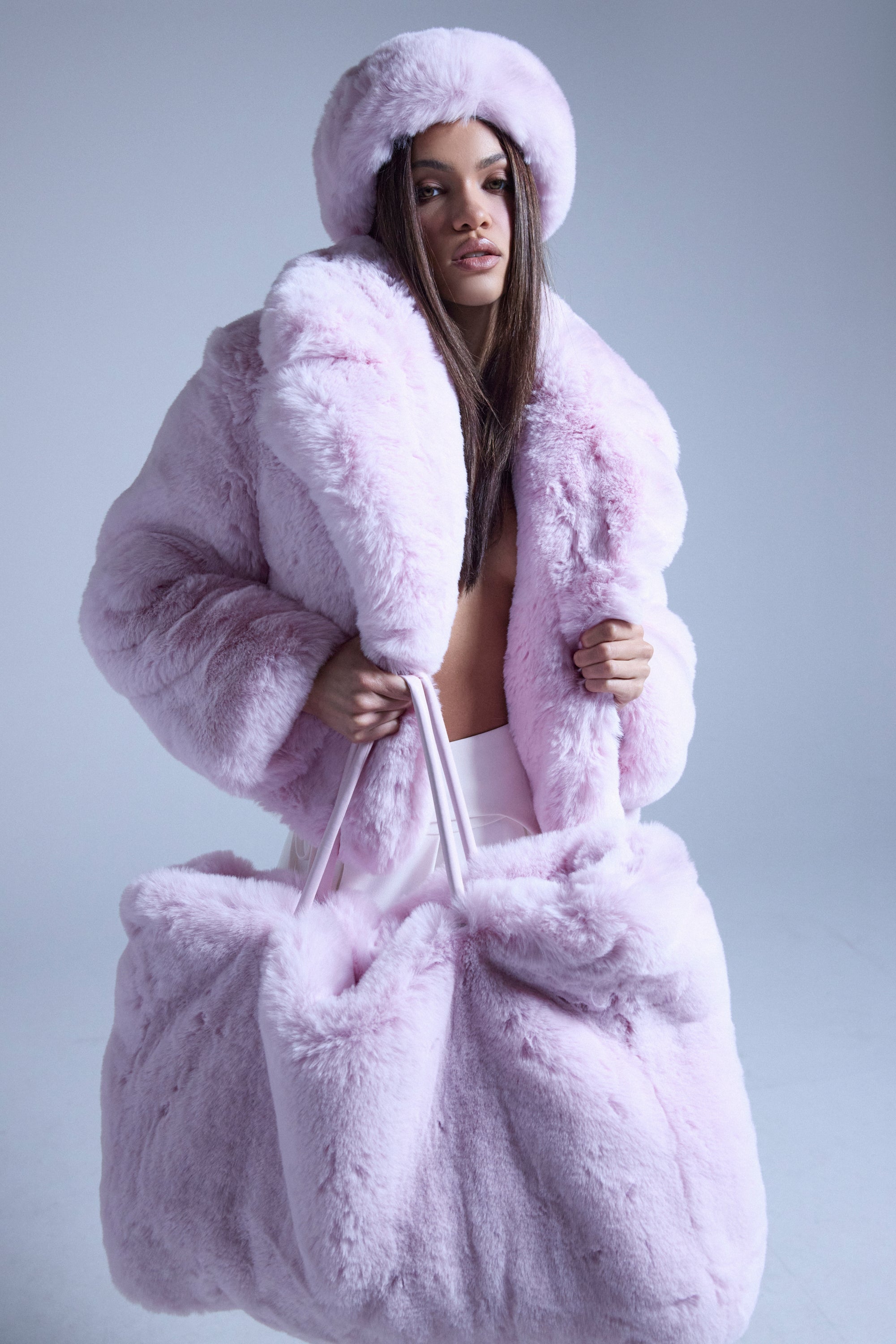 A woman wears the BBGIRL FAUX FUR COAT in pale pink with an oversized collar and matching hat, holding a large pink faux fur bag against a plain light background. 2