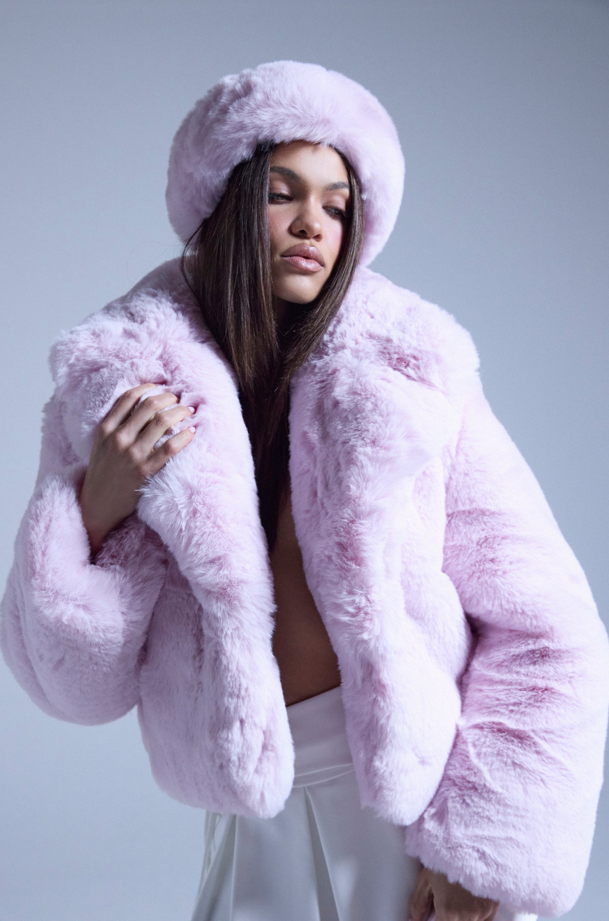 A woman poses confidently in the BBGIRL FAUX FUR COAT, featuring pink faux fur with an oversized collar and matching hat, eyes closed and dark hair visible against a plain light background. 0