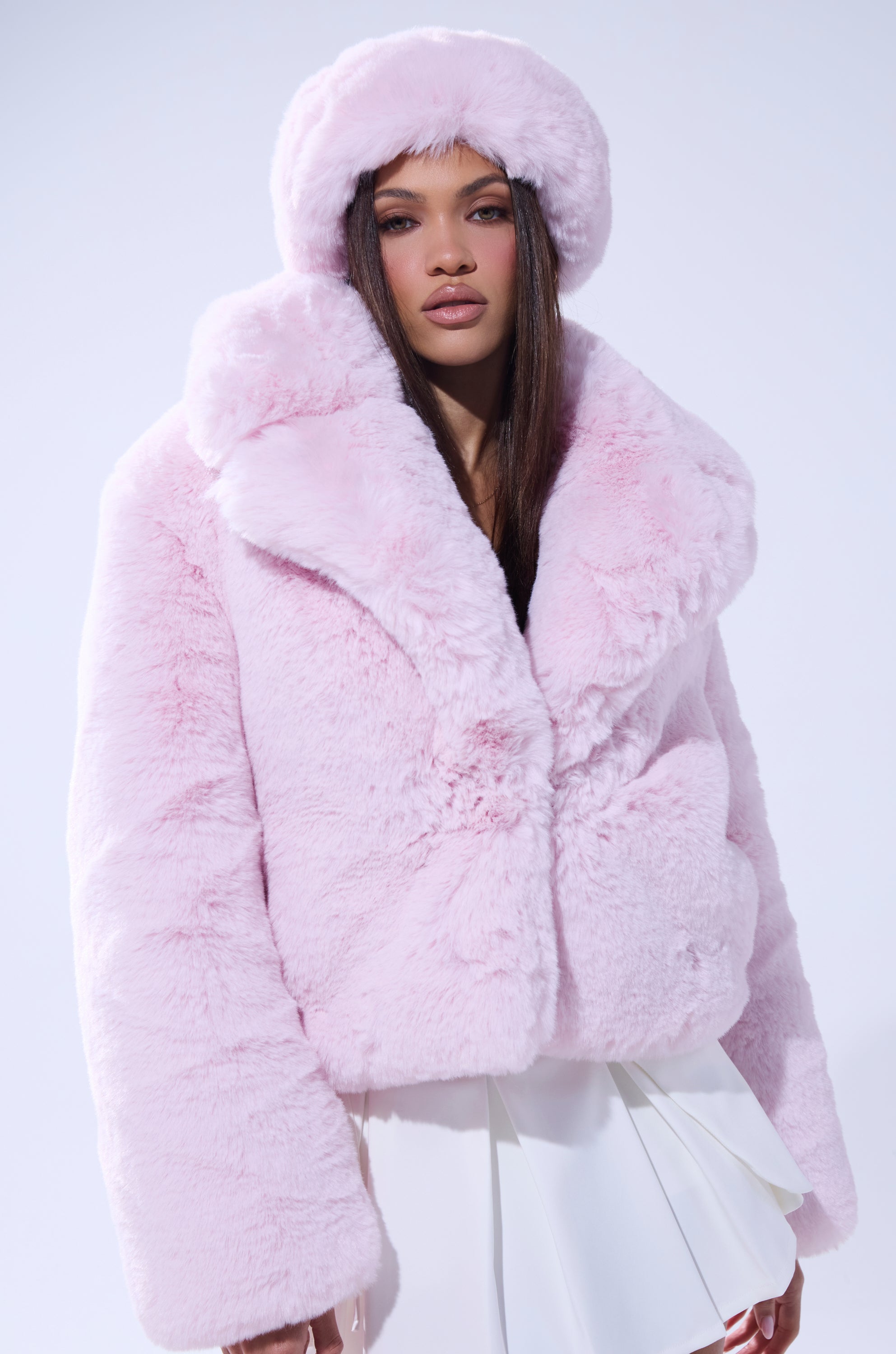 A woman poses against a white background wearing the BBGIRL FAUX FUR COAT in pink with an oversized collar, a matching hat, and a white pleated skirt. She has long straight hair. 3