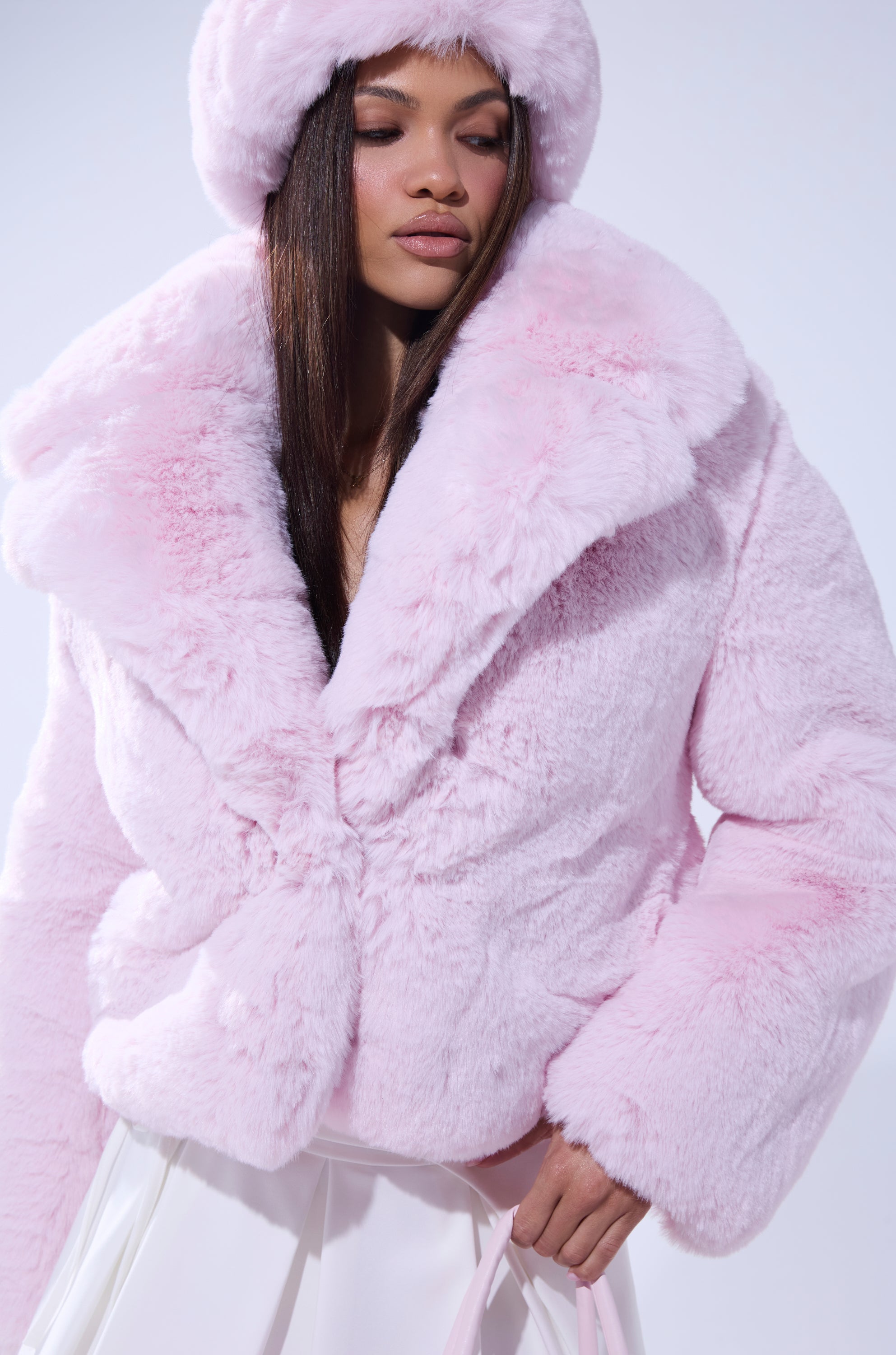Wearing the BBGIRL FAUX FUR COAT in light pink with an oversized collar and matching hat, a woman stands against a pale background holding a light pink handbag. Her long hair is straight, and she maintains a composed expression. 4