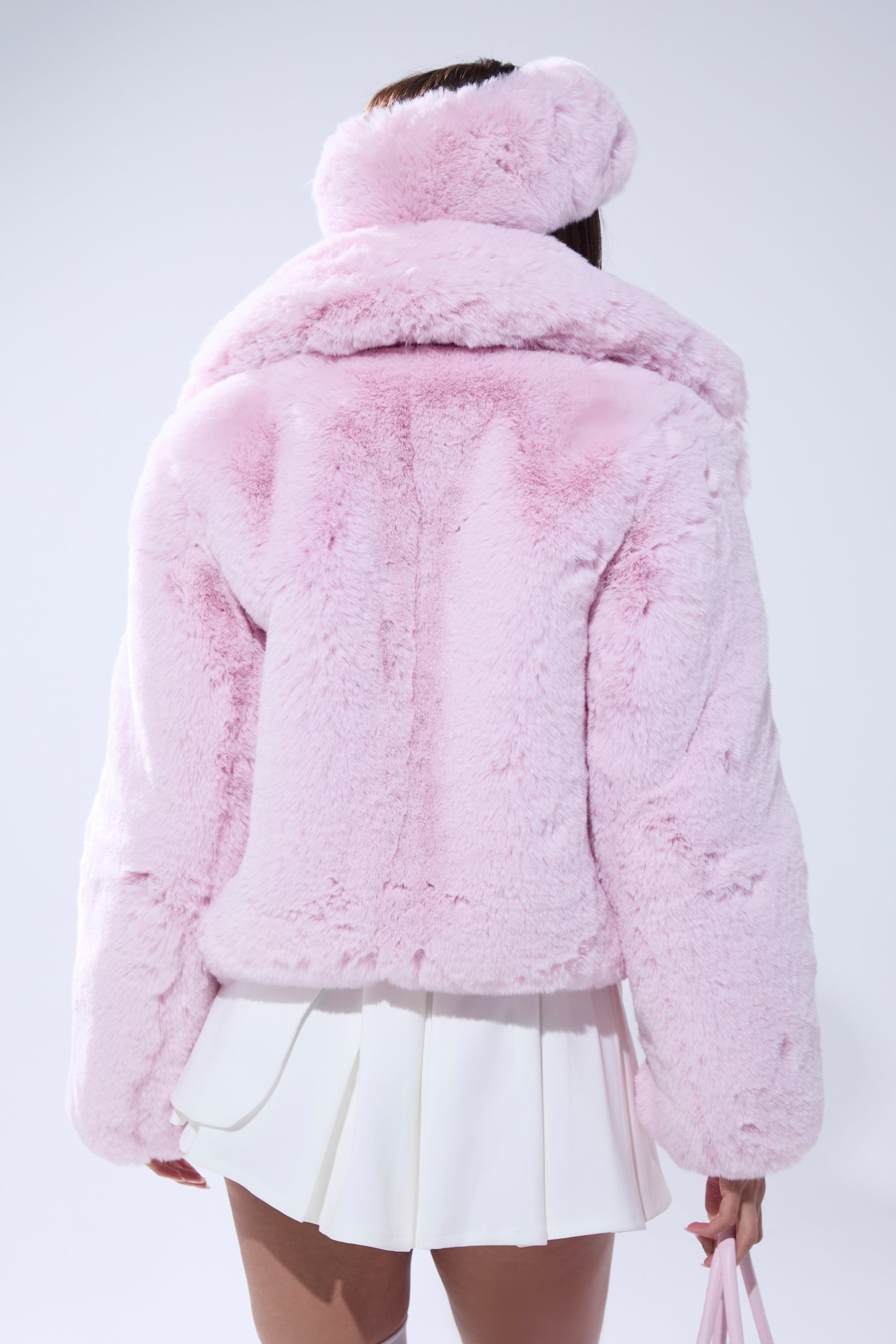 A person is seen from behind wearing the BBGIRL FAUX FUR COAT in pink with an oversized collar and matching hat, paired with a white pleated skirt and holding a pink handbag against a plain background. 5