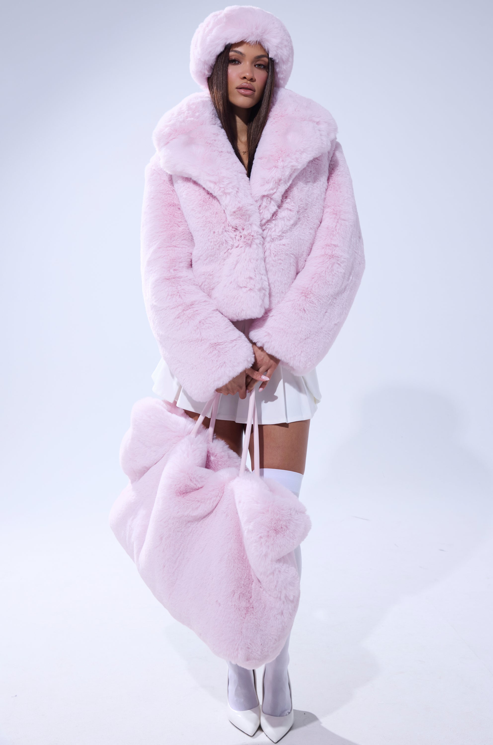 A woman models the BBGIRL FAUX FUR COAT in pink with an oversized collar and matching hat, holding a large soft pink bag. She pairs it with a white skirt, knee-high socks, and shoes against a plain light background. 6