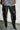 Someone stands against a white brick wall and gray concrete floor, wearing AZALEA WANG BACKER BLACK SHINE OVER THE KNEE BOOTS with stiletto heels and a sequined black-and-white patterned skirt.