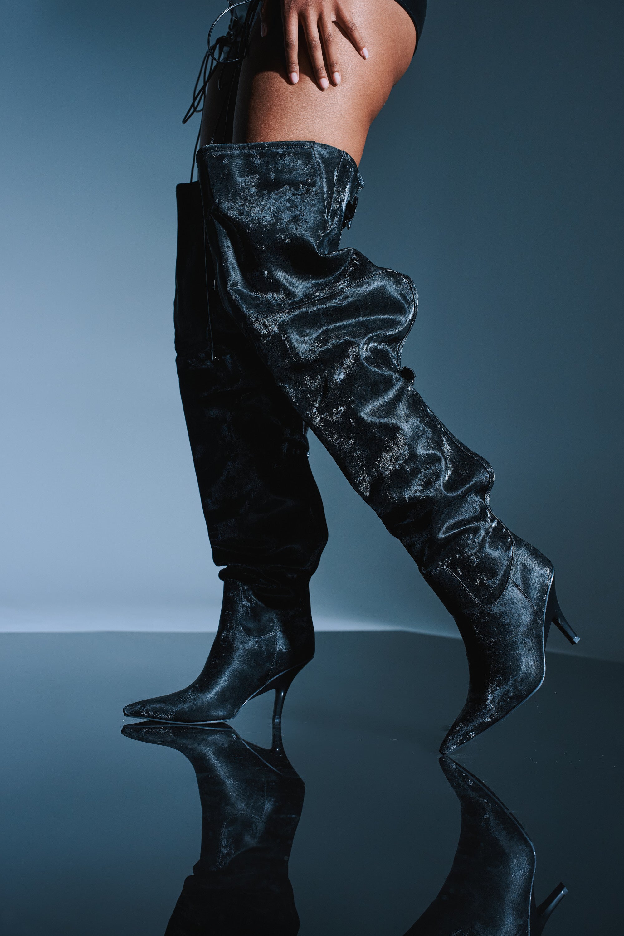 A person stands against a dark reflective surface, one hand on their thigh, wearing the AZALEA WANG BACKER BLACK SHINE OVER THE KNEE BOOT—slouchy black faux leather boots with pointed toes and stiletto heels.