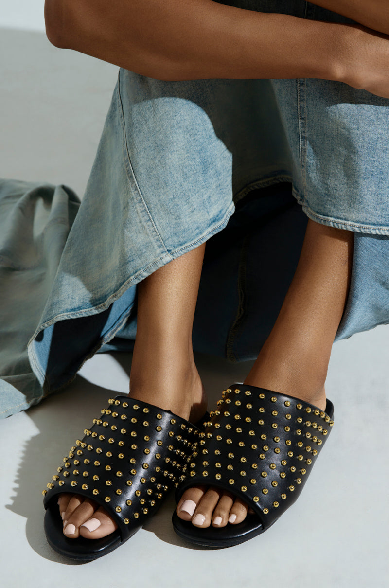 A person in a long denim skirt sits with crossed legs, showcasing the AKIRA Bagger Studded Slip-On Sandal in Black. The background is light and neutral.