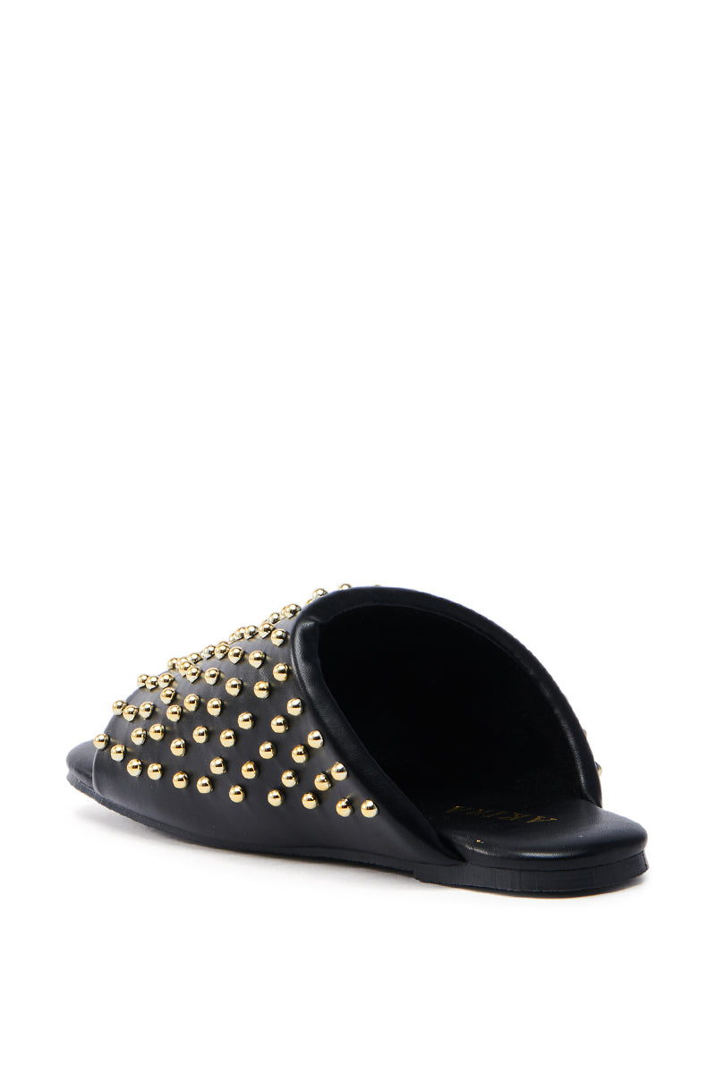 The AKIRA BAGGER Studded Slip-On Sandal in Black is made from faux leather, featuring numerous small gold studs on the upper part for a textured look. This chic sandal has a flat sole and is shown from the back at an angle over a plain white background.