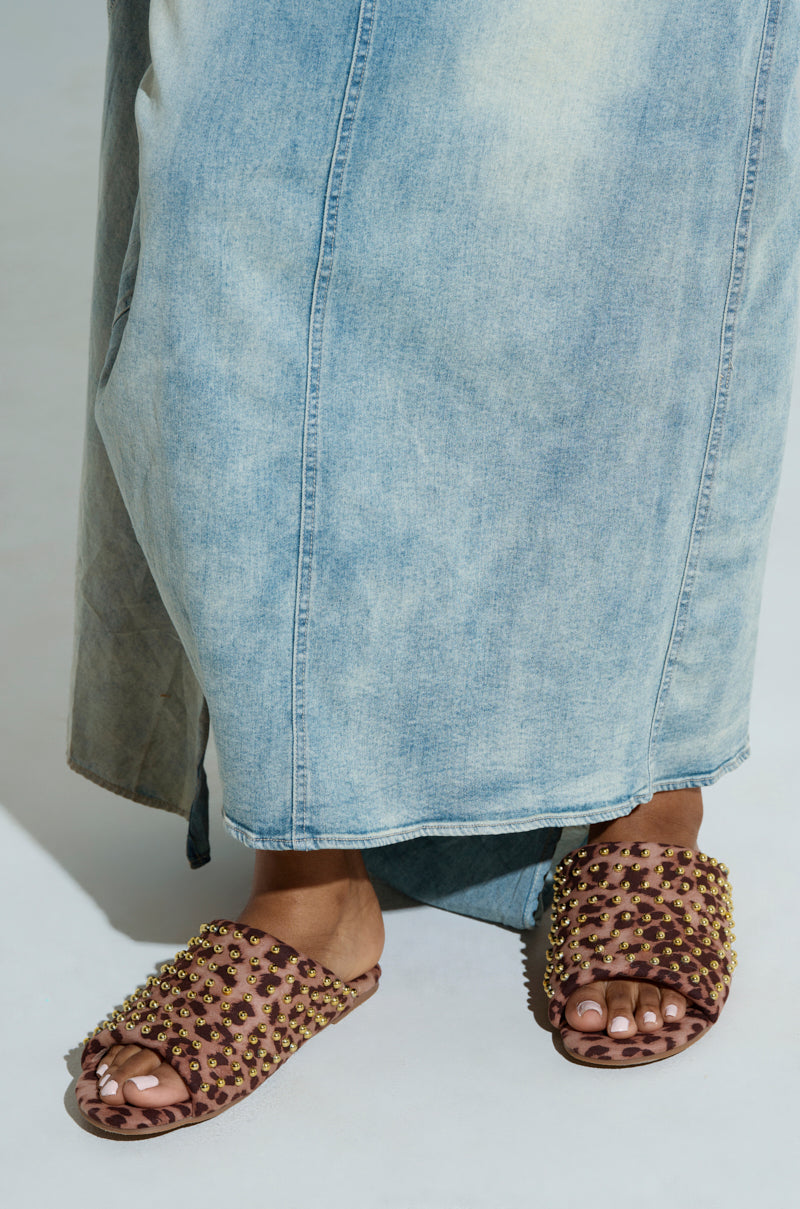A person wearing a long, light blue denim skirt stands on a light surface adorned in AKIRA BAGGER STUDDED SLIP ON SANDALS IN LEOPARD with gold-colored studs and flat rubber soles, adding flair to the ensemble.