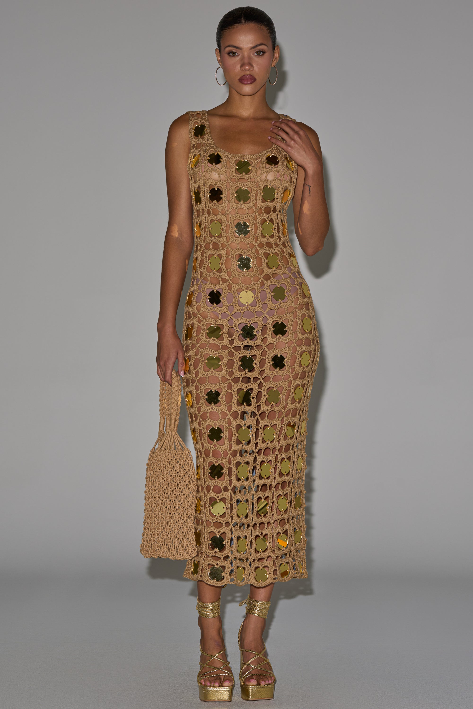 A woman poses against a plain background wearing the HOT GIRL AGENDA SEQUIN CROCHET MIDI DRESS—sleeveless, beige, with floral patterns and gold disc embellishments—styled with gold platform heels, hoop earrings, and a matching crochet bag. 0