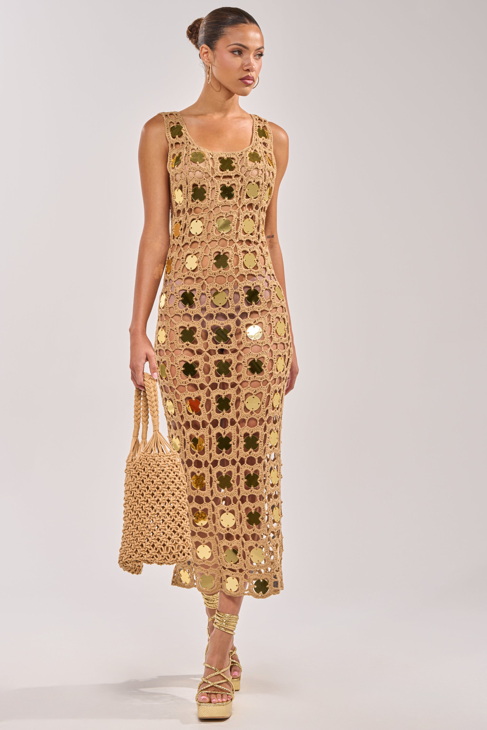 A woman wears the HOT GIRL AGENDA SEQUIN CROCHET MIDI DRESS—sleeveless, tan, with crochet floral patterns and gold sequin details. She holds a matching bag, hair in a bun, strappy sandals on, against a plain light background. 1