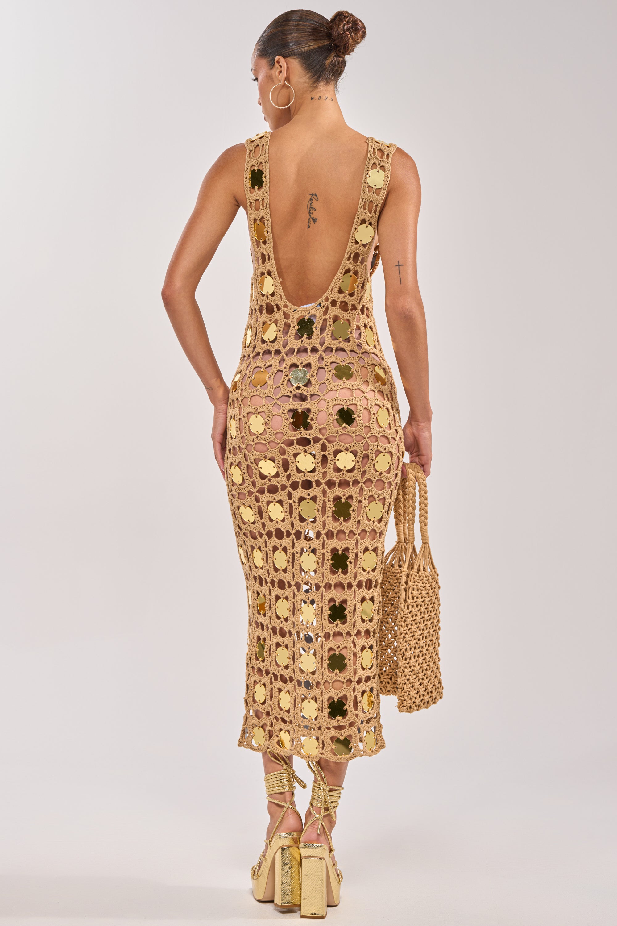 A woman with her hair in a bun wears the HOT GIRL AGENDA SEQUIN CROCHET MIDI DRESS, paired with gold platform heels and a woven handbag. She has tattoos on her upper back and right arm. 3