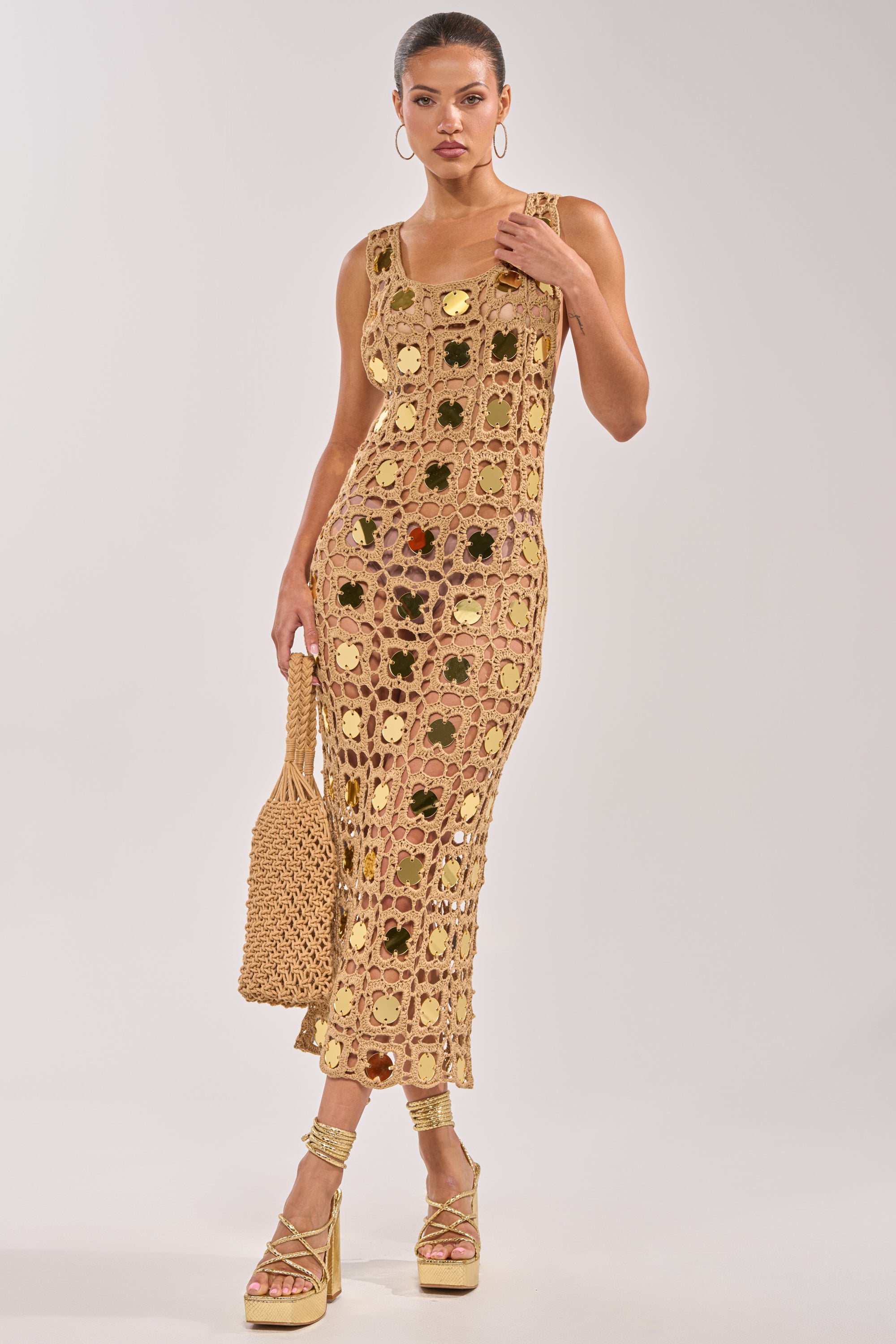 A woman wearing the HOT GIRL AGENDA SEQUIN CROCHET MIDI DRESS with gold platform heels, hoop earrings, and a woven bag stands against a plain light background. The sleeveless dress features flower cutouts and mirrored disc embellishments. 4