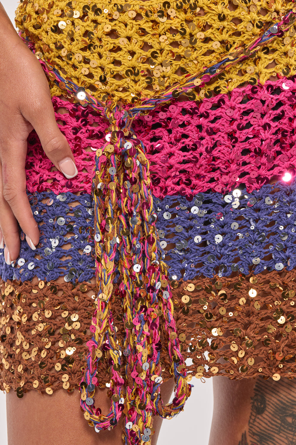 Close-up of a hand on the FULL OF LIFE CROCHET MINI SKIRT, showcasing horizontal yellow, pink, blue, and brown stripes with shiny sequins and a braided multicolored belt. Ideal for standout festival partywear style. 7