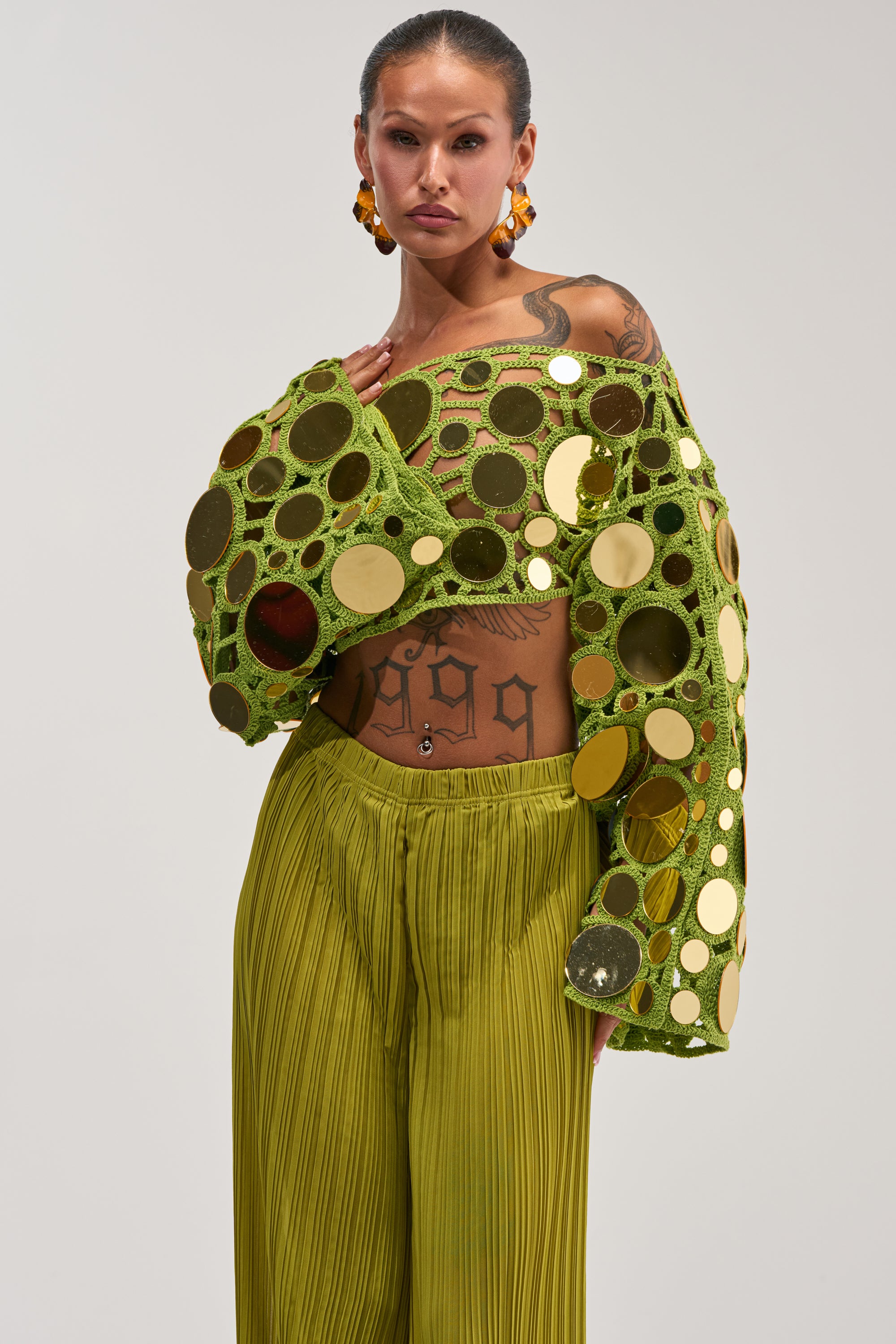 A person poses confidently against a plain background, showcasing the RESORT VIBES CROCHET CROP LONG SLEEVE IN CHARTREUSE paired with green pleated pants. The vibrant off-the-shoulder top features bold gold and green circular accents, complemented by striking earrings and visible tattoos—an ideal vacation-ready look.