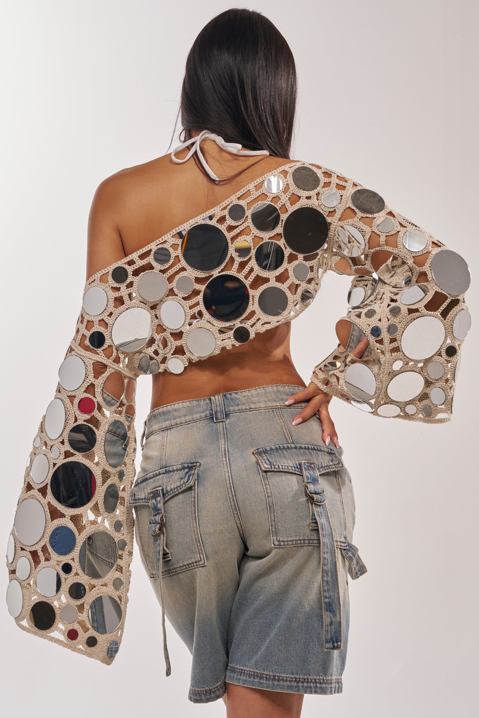 Sporting the RESORT VIBES CROCHET CROP LONG SLEEVE, a person with long dark hair and denim shorts poses with their back to the camera, showcasing the beach-ready open-weave style against a plain background. 4