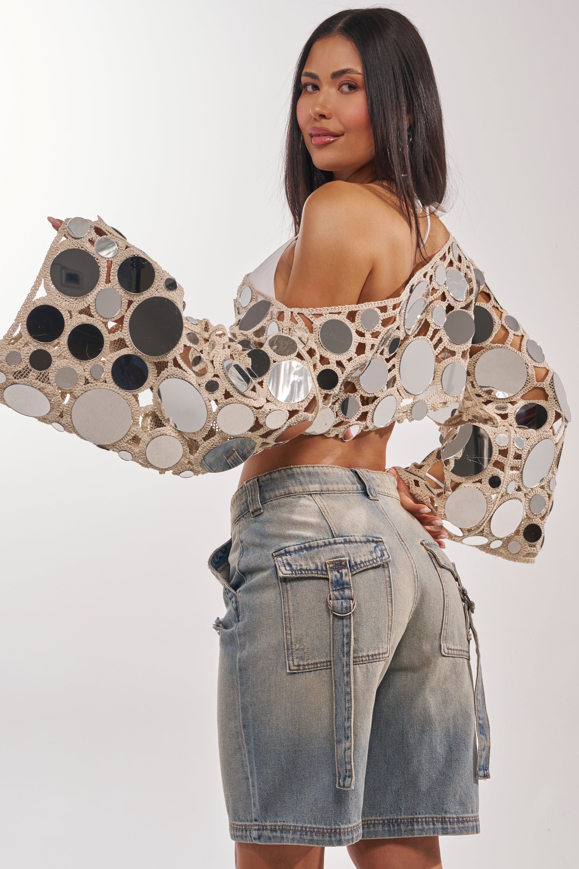 A woman with long dark hair poses in the RESORT VIBES CROCHET CROP LONG SLEEVE and light denim shorts, creating a chic, modern resort look that's perfect for the beach against a plain background. 5
