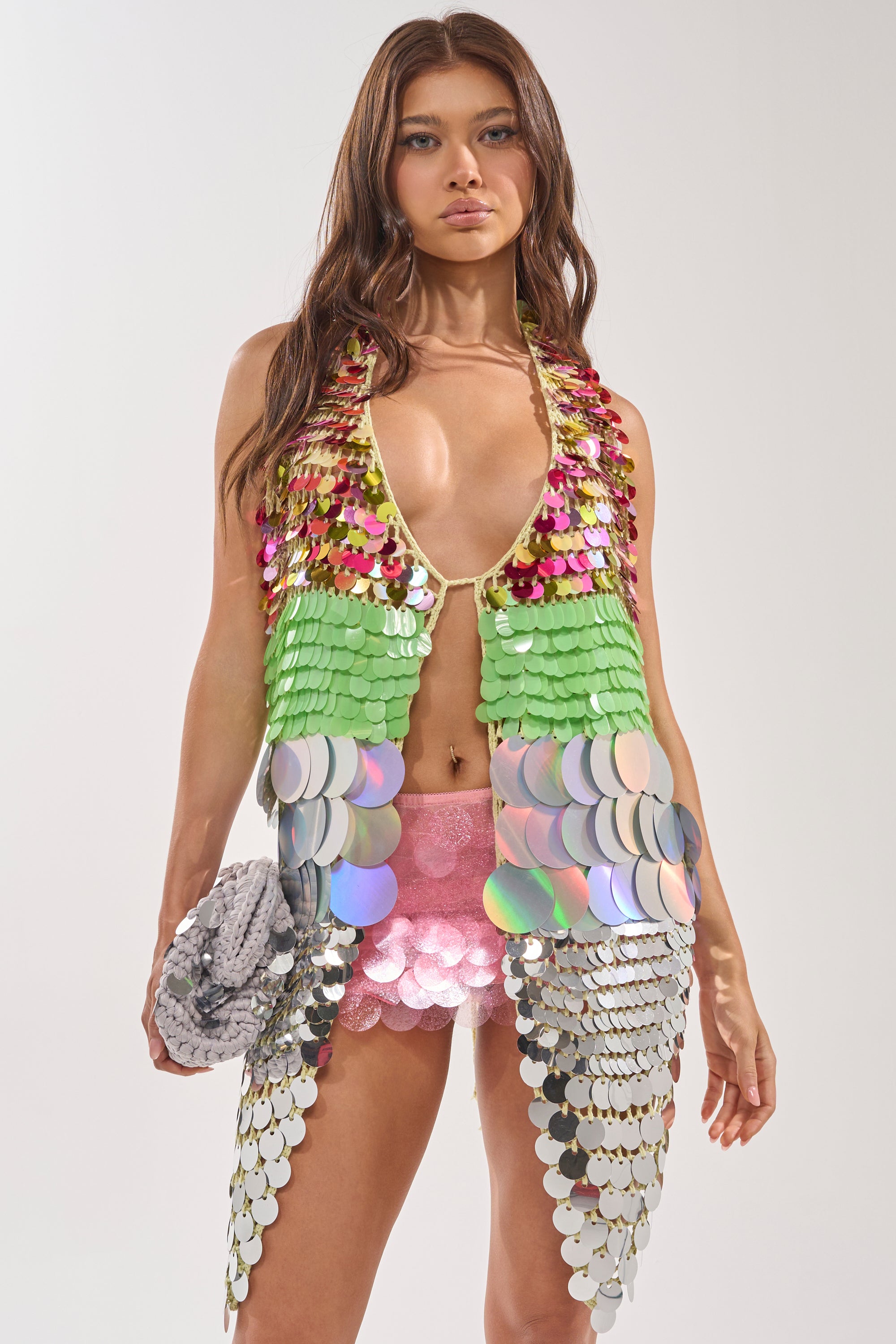 A woman with long brown hair wears the SHAKE IT GIRL CROCHET SEQUIN TOP—a colorful green, gold, and silver festival vest—paired with pink sequined shorts and a silver clutch, posing against a plain light backdrop. 0