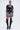 A woman stands confidently in the BOUJEE BANDANA SEQUIN MINI DRESS IN BLACK, styled with layered necklaces, large hoop earrings, and knee-high black patent boots featuring multiple buckles.