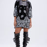 A woman stands confidently in the BOUJEE BANDANA SEQUIN MINI DRESS IN BLACK, styled with layered necklaces, large hoop earrings, and knee-high black patent boots featuring multiple buckles.