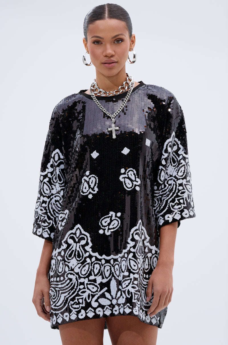 A woman wears the BOUJEE BANDANA SEQUIN MINI DRESS IN BLACK—a loose-fitting mini dress with white bandana patterns and half-length sleeves—paired with layered silver necklaces, including a cross pendant, against a plain light background.