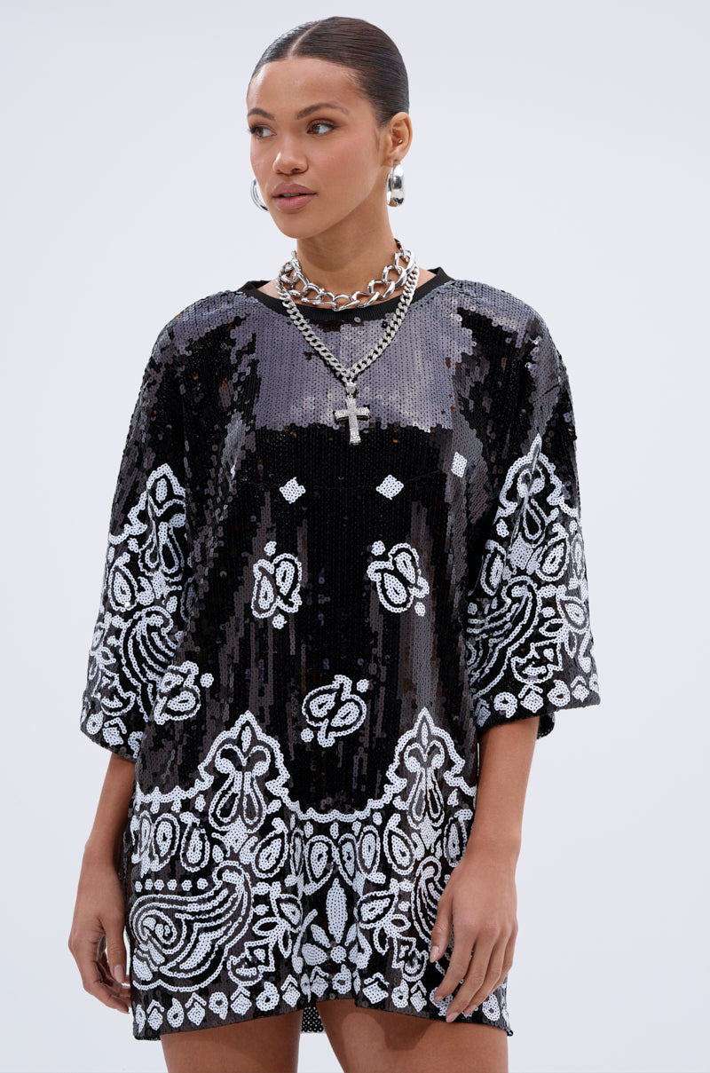 A woman stands against a plain light background wearing the BOUJEE BANDANA SEQUIN MINI DRESS IN BLACK, featuring a loose fit with intricate white patterns, styled with chunky silver necklaces and a cross pendant.