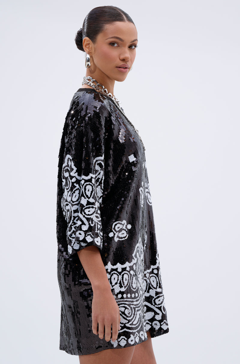 A woman with slicked-back hair models the BOUJEE BANDANA SEQUIN MINI DRESS IN BLACK, featuring black and white ornate sequins, as she stands sideways and looks at the camera against a plain light background.