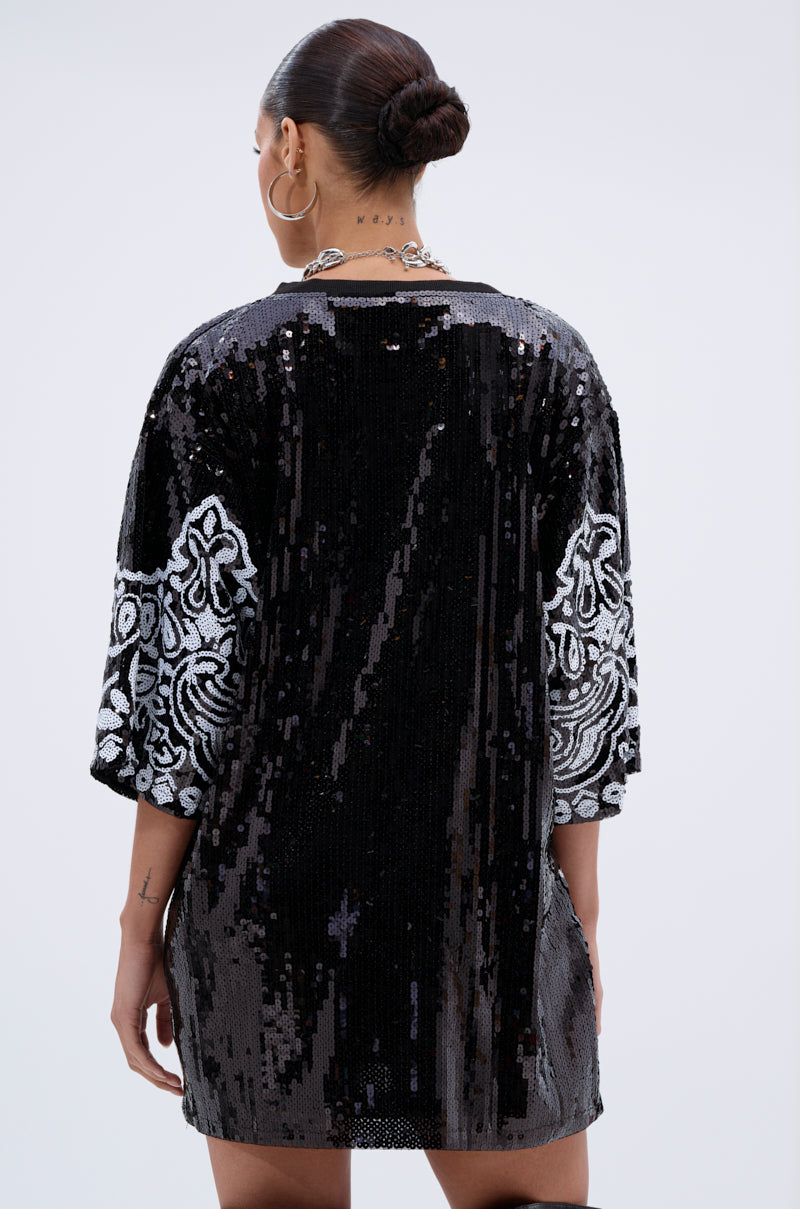 A person faces away, wearing the BOUJEE BANDANA SEQUIN MINI DRESS IN BLACK with white ornate sequin designs on the sleeves. Their hair is in a sleek bun, finished with large hoop earrings and a silver chain necklace.