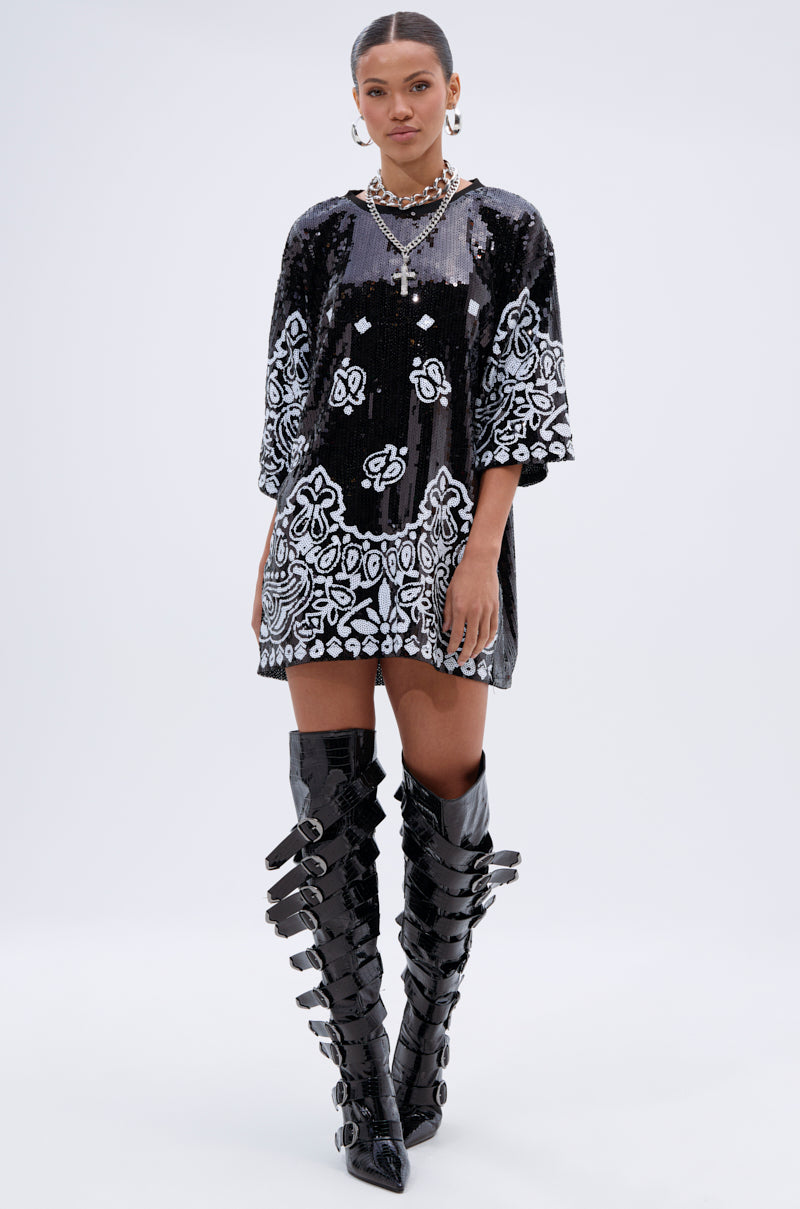 A woman wears the BOUJEE BANDANA SEQUIN MINI DRESS IN BLACK, featuring white ornate patterns, with knee-high black buckle boots, chunky necklaces, and hoop earrings against a plain light background.