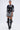 A woman wears the BOUJEE BANDANA SEQUIN MINI DRESS IN BLACK, featuring white ornate patterns, with knee-high black buckle boots, chunky necklaces, and hoop earrings against a plain light background.