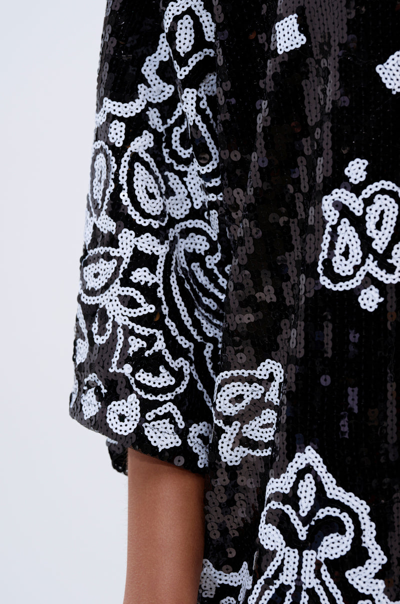 Close-up of a person's arm in the BOUJEE BANDANA SEQUIN MINI DRESS IN BLACK, showcasing intricate white sequin and floral patterns on an elbow-length sleeve, set against a plain, light-colored background.