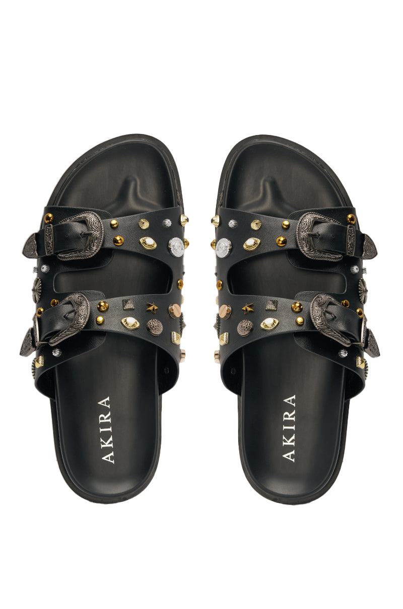 The AKIRA BARCLAY BLACK METAL EMBELLISHED SLIP ON FLAT SANDAL features black faux leather, two buckled straps adorned with gold, silver, and gem-like studs, and "AKIRA" in white on the insoles. 3