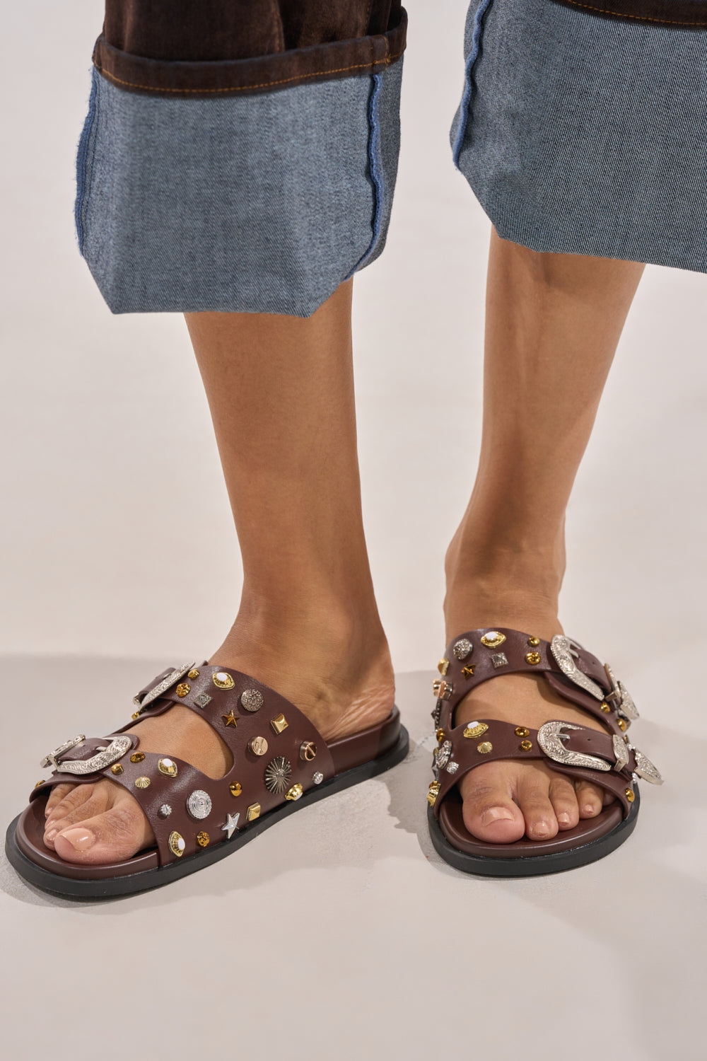 Wearing wide-cuffed blue jeans, the person sports the AKIRA BARCLAY BROWN METAL EMBELLISHED SLIP ON FLAT SANDAL, featuring silver and gold studs and large ornate buckles. The photo spotlights their feet and lower legs. 1