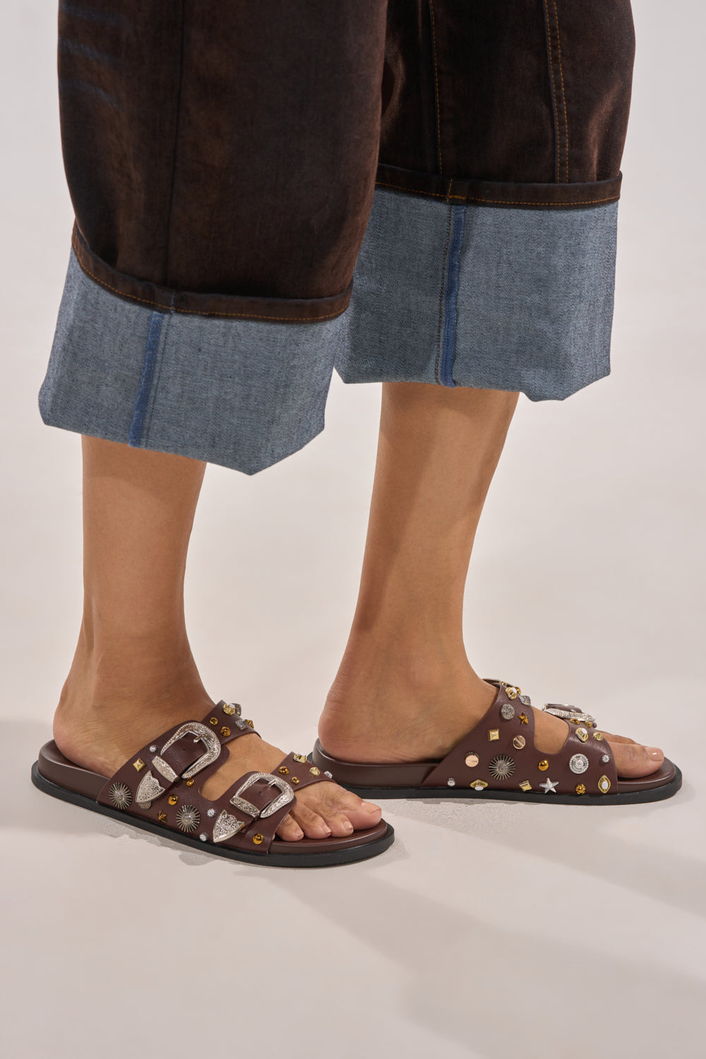 Wearing dark cuffed denim pants and AKIRA BARCLAY Brown Metal Embellished Slip On Flat Sandals, a person stands on a light-colored surface. 2