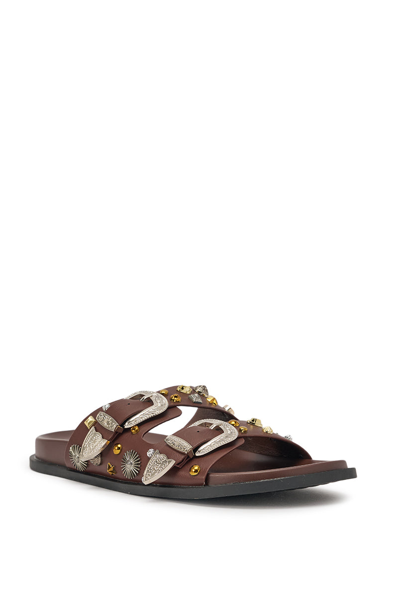 The AKIRA BARCLAY BROWN METAL EMBELLISHED SLIP ON FLAT SANDAL features two wide brown faux leather straps with large silver buckles, metallic gold and silver studs, and a flat black low-profile sole. 5