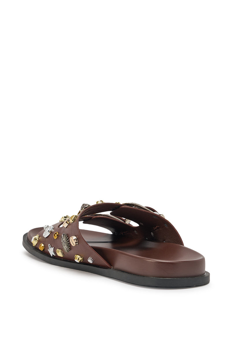 The AKIRA BARCLAY Brown Metal Embellished Slip On Flat Sandal features brown faux leather and a black sole, adorned with gold and silver metal studs in various shapes. The sandal is pictured from the rear side angle. 6