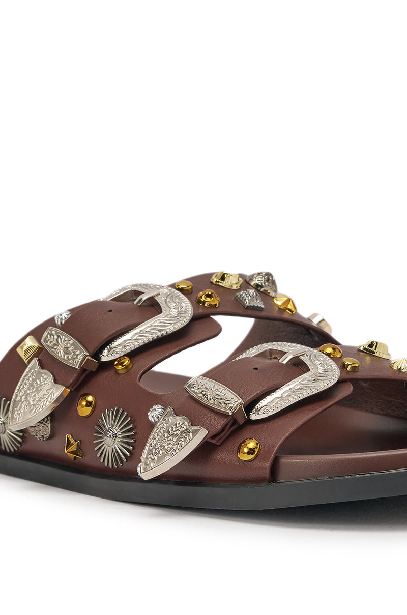 The AKIRA BARCLAY BROWN METAL EMBELLISHED SLIP ON FLAT SANDAL features a brown design with two straps, ornate silver buckles, and gold and silver metal studs on a black sole. 7